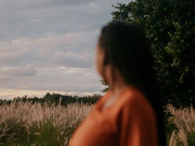 A Black woman with long hair is wearing an orange dress is look away toward the field behind her.